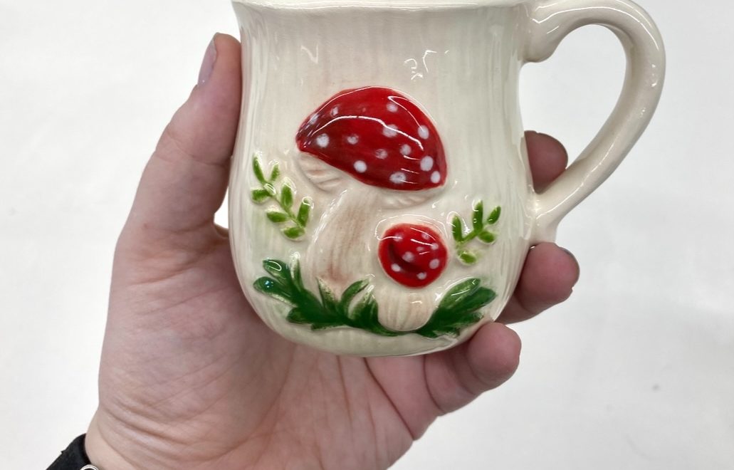 Finished ceramic mug with red and white mushrooms and green plants on a white background with shiny finish.